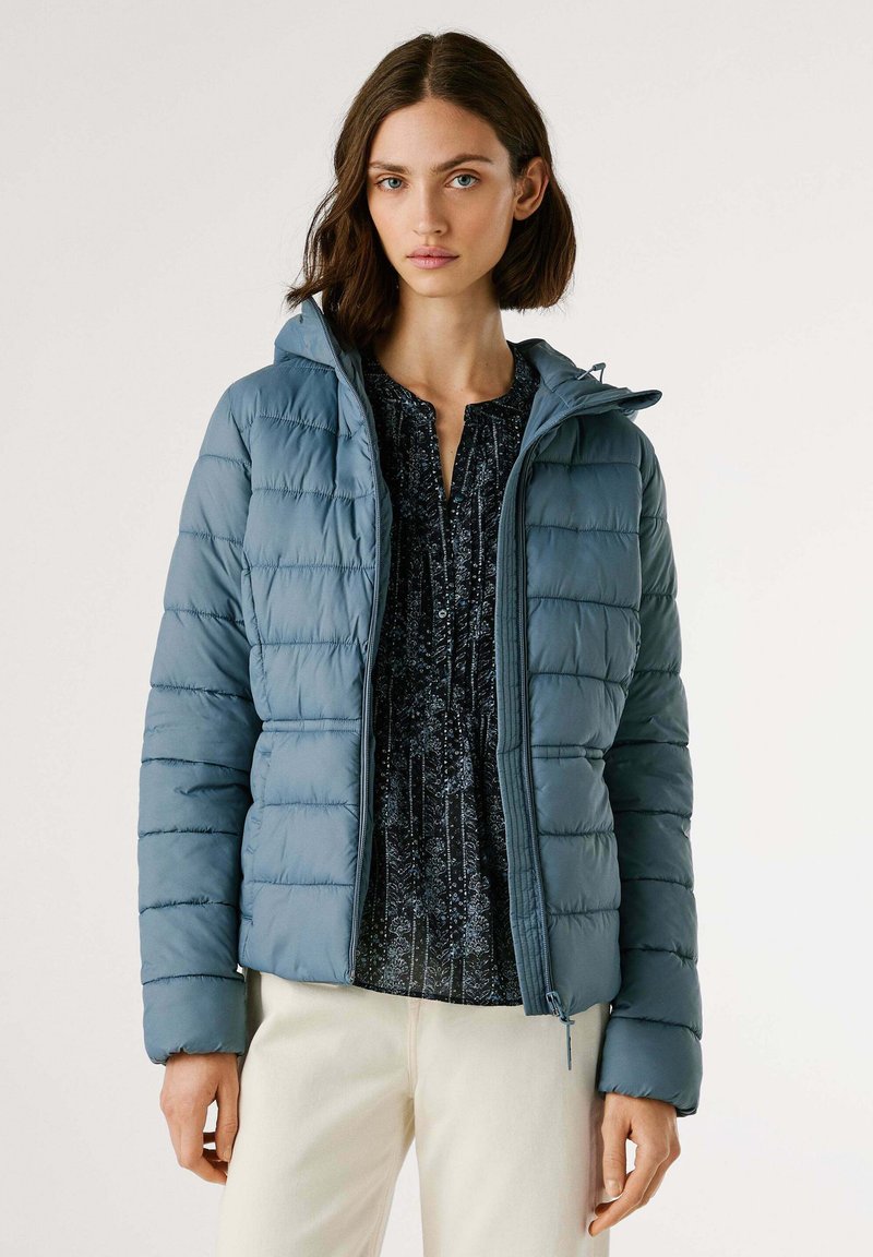 Vrouw in een blauwe gewatteerde jas met capuchon, over een donker gemarmerde blouse en lichtgekleurde broek, staande tegen een effen achtergrond.