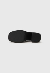 Suela negra de zapato con patrón de agarre acanalado, tacón rectangular y área del arco lisa sobre fondo blanco liso.