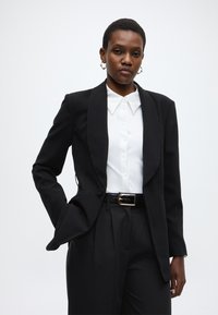 Μαύρο σακάκι με σατέν πέτο, λευκό πουκάμισο με κουμπιά, μαύρο παντελόνι ψηλόμεσο και υφασμένη μα ύρη ζώνη με χρυσή αγκράφα.