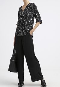 Schwarze Bluse mit langen Ärmeln und weißem Sternenmuster; V-Ausschnitt-Design. Kombiniert mit schwarzen, weiten Hosen und einer schwarzen Handtasche.