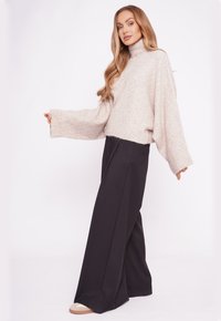 Kobieta w beżowym, oversize'owym swetrze z wysokim kołnierzem, szerokich czarnych spodniach i beżowych sneakersach, stojąca na tle gładkiej białej ściany.