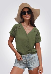 Camisa de botón en verde oliva con tejido texturizado y acentos de encaje en las mangas, combinada con shorts de mezclilla desgastados en azul claro. Sombrero de ala ancha.