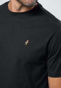 Artimos planas vyro, vilkinčio juodą marškinėlį su apvalia apykakle, kurio krūtinėje yra maža auksinė išsiuvinėta žmogaus figūra, einanti.