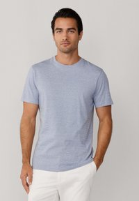 Uomo con capelli scuri, che indossa una maglietta a maniche corte azzurro chiaro e pantaloni bianchi, in piedi davanti a uno sfondo chiaro e semplice.