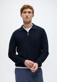 Σκούρο ναυτικό μπλουζάκι με γιακά, ριμπ υφή και φερμουάρ στο ένα τέταρτο. Φορεμένο πάνω από λευκό πουκάμισο, διαθέτει μακριά μανίκια και εφαρμοστά μανσέτες.