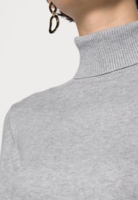 Ljusgrå polokrage med ribbad textur, med hög krage. Åtföljd av en guldfärgad cirkulär örhänge med en strukturerad yta.