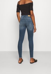 Jeans ajustados de color azul oscuro con una cintura alta, con un ligero desgaste y dos bolsillos en la parte posterior. Se llevan con una blusa negra caída sobre los hombros y sandalias plans.