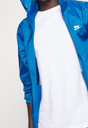 Person iført en knaldblå Nike lynlåsjakke over en plain hvid T-shirt, jakken er delvist åbnet og hætten er oppe.