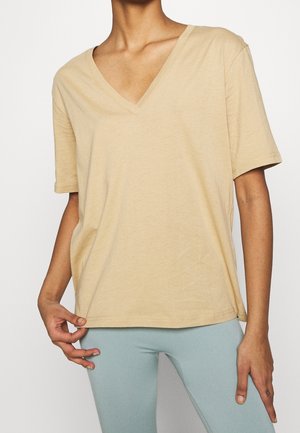 Persoon die een loszittend beige T-shirt met korte mouwen en V-hals draagt en lichtblauwe legging, het shirtzoom met één hand vasthoudend.