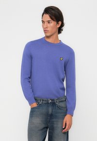Lila Strickpullover mit rundem Ausschnitt, langen Ärmeln und einem kleinen gelben Vogel-Logo auf der linken Brust. Tragen mit ausgewaschenen blauen Jeans.