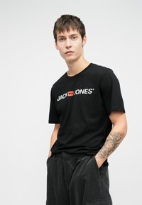 Černé bavlněné tričko s bílým a červeným logem "JACK & JONES". Model má na sobě volné tmavě šedé manšestrové kalhoty, na jeho ruce je viditelné tetování.
