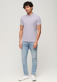 Lichtpaarse polo met kraag, korte mouwen en geborduurd logo, gecombineerd met lichtblauwe versleten jeans en witte sneakers.