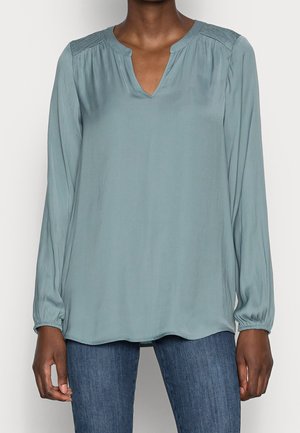 Femme portant un chemisier bleu clair à manches longues avec un col en V et des épaules froncées, associé à un jean bleu moyen délavé.