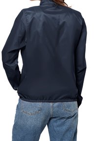 Marinblå lättviktsjacka med en slät, glänsande yta, hög krage och elastisk fåll, bärs över ljusblå jeans. Bakifrån sett.