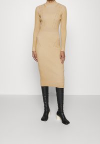 Vestito a maglia in beige chiaro con righe verticali, maniche lunghe e collo alto; abbinato a stivali neri sopra il ginocchio con tacchi bianchi.