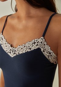 Camisole marine avec des bordures en dentelle beige festonnées le long de l'encolure, fines bretelles et une silhouette ajustée. Texture de tissu lisse.