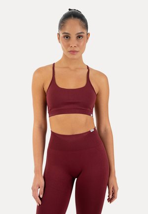 Fit vrouw met donker haar in een knot draagt een bordeauxrode sport-bh en hoge taille legging tegen een effen lichte achtergrond.