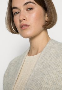 Šviesiai pilkas, tekstūruotas vilnos drabužis su plačiu apykakle, uždėtas ant kreminių spalvų megztinio su vertikaliu raštu.