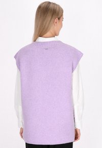 Lila mouwloze trui met geribbelde halslijn, zachte textuur en een klein logo-detail op de achterkant. Gepaard met een witte overhemd met kraag eronder.