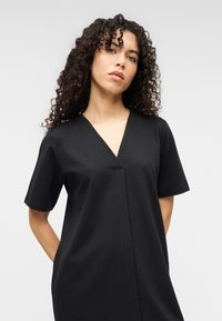 Μαύρο φόρεμα με κοντά μανίκια, με V-λαιμόκοψη και καθαρές γραμμές, κατασκευασμένο από ομαλό ύφασμα με διακριτική λάμψη. Απλός και μοντέρνος σχεδιασμός.