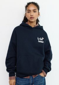 Felpa con cappuccio blu navy con tasca frontale, cappuccio con cordoncino e logo bianco "Le Chef Lorenzo". Indossata sopra una camicia a quadri.