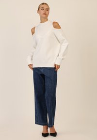 Biała bluza z odkrytymi ramionami, tekstura prążkowana przy dekolcie, w połączeniu z niebieskimi spodniami typu wide-leg z drobnymi dekoracyjnymi akcentami.