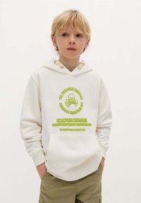 Crème hoodie met groen geborduurd doodskop- en gekruiste bottenlogo, verstelbare nekstrikjes, zachte stof en een voorzak. Geraadpleegd met olijfgroene shorts.