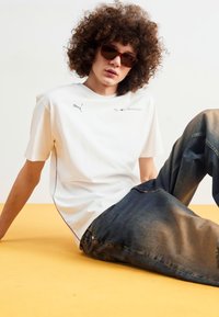 Camiseta de algodón blanca con costuras negras y logo de Puma, combinada con vaqueros de mezclilla azules con textura desgastada. Gafas de sol marrones puestas.