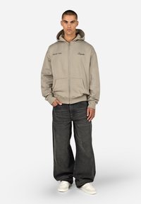 Felpa con zip color beige dotata di tasche frontali e ricamo "Drivers Club". Abbinata a jeans scuri a vestibilità ampia e sneakers bianche.