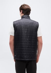 Mees, kes kannab musta tikanditega varrukateta vest valge t-särgi peal, seisab seljaga, taust on ühtlaselt valge.