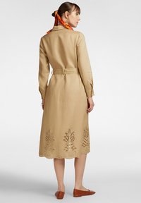 Abito in lino beige con maniche lunghe, vita con cintura e ricami decorativi a forma di taglio sull'orlo. Le scarpe marroni senza lacci completano il look.