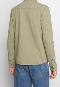 Khaki košile s dlouhým rukávem, s límečkem, knoflíkovými manžetami a hladkým povrchem. Spárována s modrými džínami s kapsami na zadní straně.