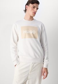 Weißer Pullover mit einem beigefarbenen rechteckigen Grafikdesign, auf dem das Wort "HUGO" in fetten weißen Buchstaben steht. Gerippte Bündchen und Saum, weicher Stoff.