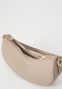 Beige textured leather Lacoste shoulder bag with gold zipper and logo, crescent shape, and adjustable strap on a white background.