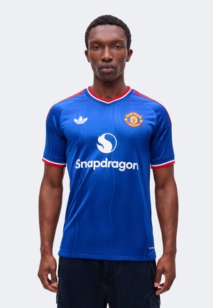 Giovane uomo che indossa la maglia da calcio blu del Manchester United con il logo Snapdragon, in piedi davanti a uno sfondo semplice, volto in avanti.