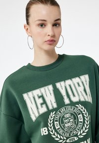 Zielona bluza z nadrukiem "NEW YORK" w wyrazistych białych literach, z emblematem uniwersytetu oraz wzorem laurowym. Miękka tekstura, okrągły dekolt.