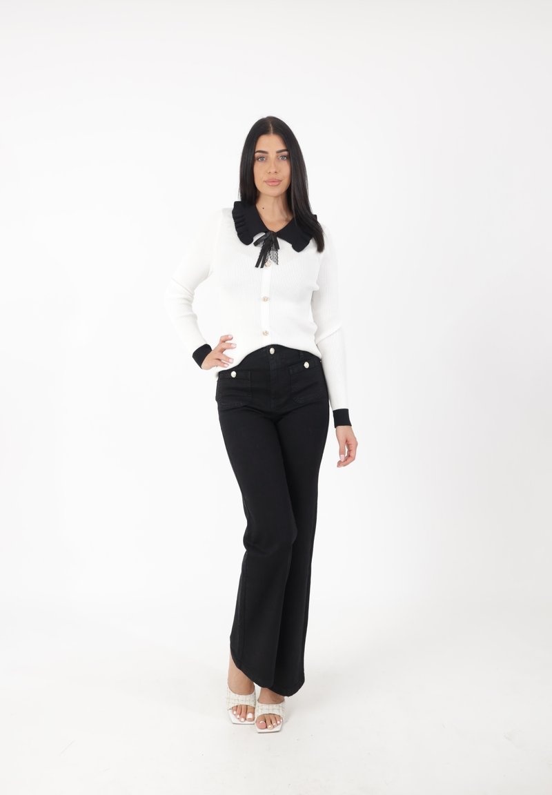 Cardigan blanc à côtes avec col à volants noir et lien en dentelle, associé à un jean noir évasé taille haute et des talons blancs à lanières.