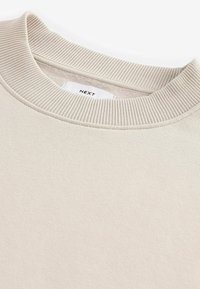 Un sweat-shirt beige avec un col ras du cou côtelé et une texture douce, portant une étiquette en tissu visible avec l'inscription "NEXT."