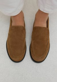 Mocassins en daim marron avec un bout rond, des coutures décoratives et une semelle en caoutchouc noir. Texture lisse et design sans lacets.