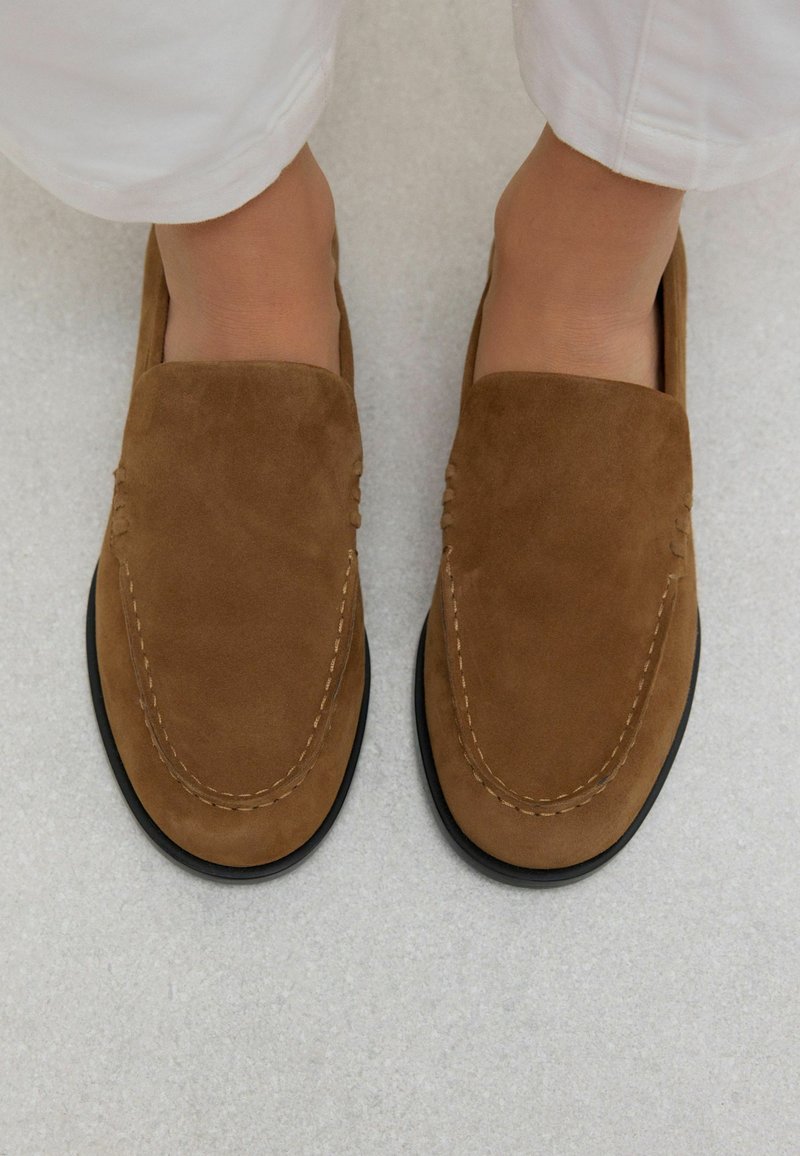 Mocassins en daim marron avec un bout rond, des coutures décoratives et une semelle en caoutchouc noir. Texture lisse et design sans lacets.