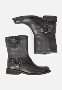 Botas de cuero negras hasta el tobillo con punta redonda, hebilla lateral y una correa ajustable que presenta un acento metálico circular. Suela de goma plana.