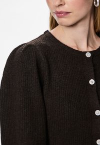 Femeie purtând un cardigan tricotat texturat maro închis cu mâneci bufante și nasturi albi, arătând buzele, gâtul și o cercea argintie rotundă.