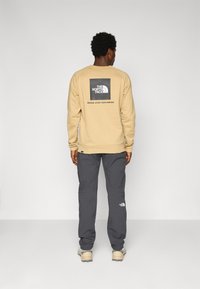 Beige sweatshirt met een zwart logo op de achterkant, gecombineerd met donkergrijze broek. Zachte stof met een relaxed fit en contrasterende merkdetails.