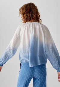 Blusa leggera e trasparente con una sfumatura ombre che va dal bianco al blu, con polsini elastici e scollatura arricciata. Abbinata a jeans blu con motivo.