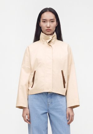 Γυναίκα με μακριά σκούρα μαλλιά, που φοράει μπεζ μπουφάν με ψηλό γιακά και καφέ φερμουάρ και ανοιχτόχρωμο μπλε τζιν, στέκεται μπροστά σε απλό φόντο.