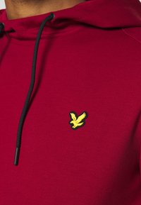 Roter Hoodie aus weichem Stoff mit Kapuze und Kordelzug. Verfügt über ein gelbes Adler-Logo im linken Brustbereich und schwarze Kordelzug-Akzente.