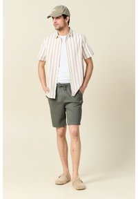 Camisa de botones de rayas claras con mangas cortas, camiseta blanca debajo, pantalones cortos grises, zapatos de slip-on beige y una gorra clara.