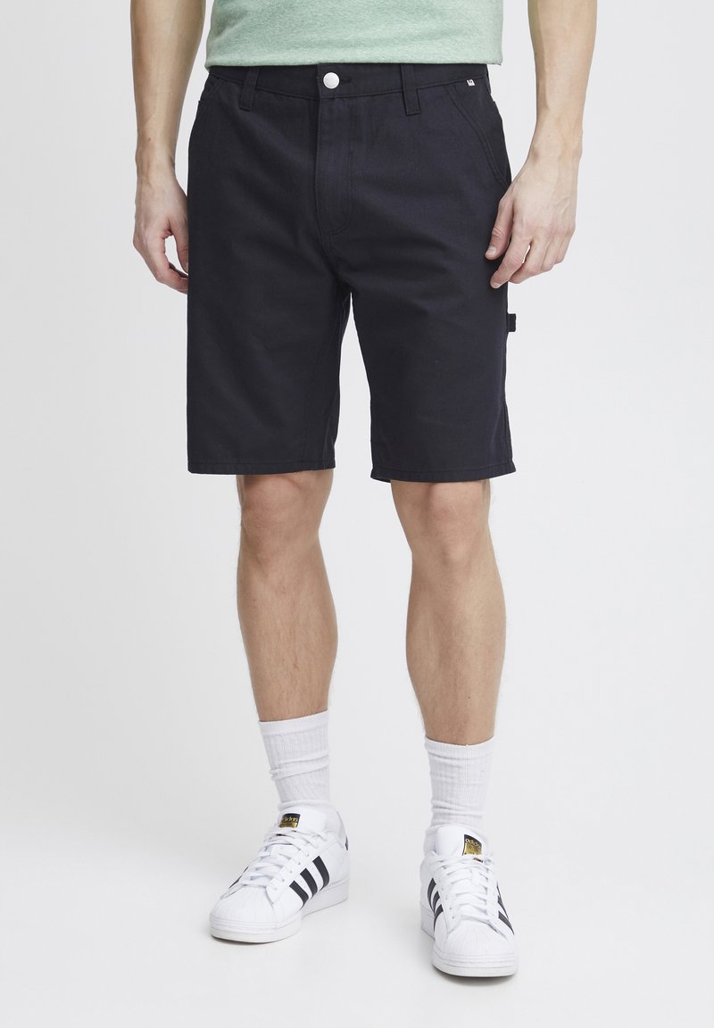 Pantalones cortos de mezclilla negra con un corte recto, bolsillos frontales y un bolsillo lateral con parche. Combinados con calcetines blancos y zapatillas blancas clásicas.