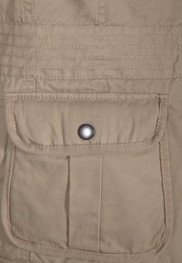 Beige tygficka med lock som är säkrad av en enda metalltryckknapp, med förstärkt sömnad och en lätt skrynklig textur.