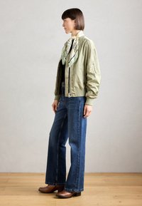 Chaqueta bomber en verde oliva claro, pantalones vaqueros azules de campana y holgados, zapatos de tacón marrones, bufanda blanca con patrón floral verde y suelo de madera.